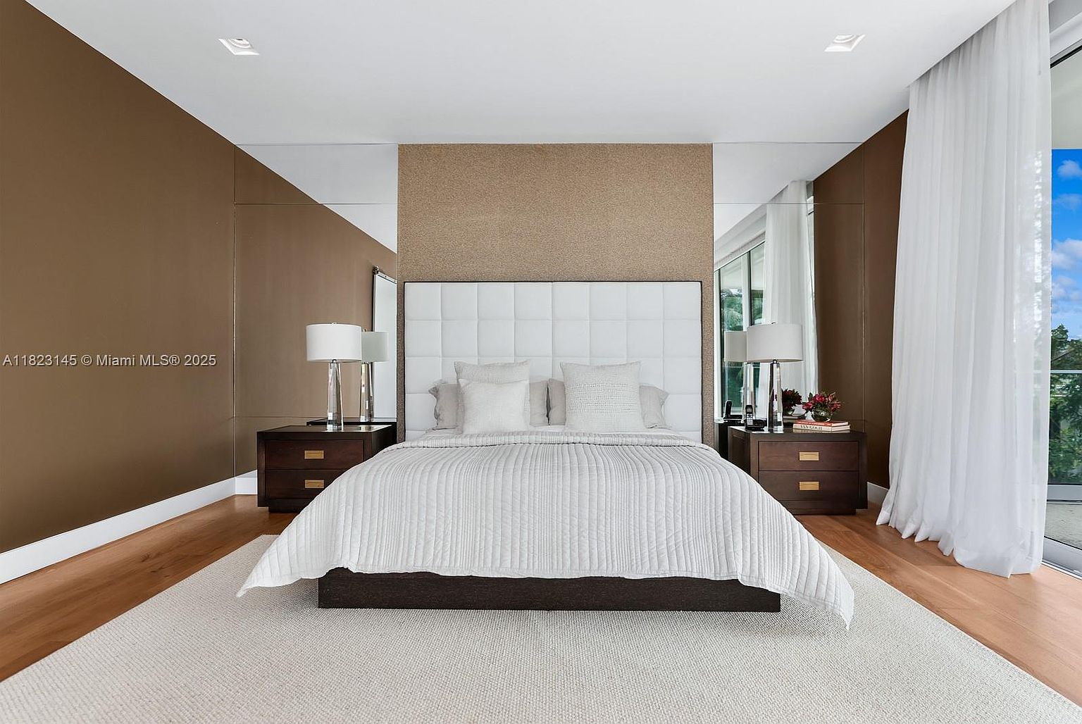 This primary bedroom features a centered bed with a large, tufted white headboard against a textured accent wall, flanked by dark wood nightstands and matching table lamps. The room is framed by mirrored wall panels on either side, reflecting the space and adding depth, while light wood flooring and a neutral area rug provide a warm foundation. To the right, floor-to-ceiling windows draped in sheer white curtains allow for natural light and a view of the outdoors.