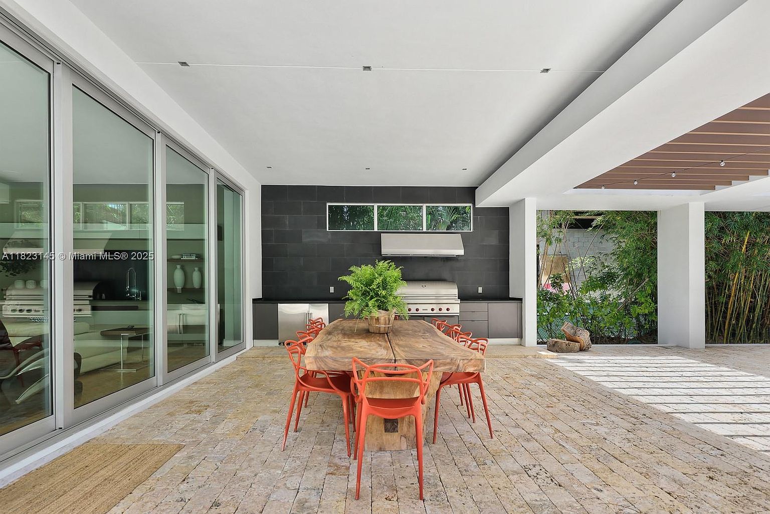 This expansive covered outdoor patio features a large, rustic live-edge wood dining table surrounded by vibrant orange chairs, creating a striking focal point. The space is equipped with a built-in outdoor kitchen featuring a grill and dark cabinetry against a dark tiled wall, while large glass sliding doors provide a seamless transition to the interior. The patio is paved with light-toned stone tiles and opens up to a lush, green backyard area, offering a sophisticated and airy setting for outdoor entertaining.