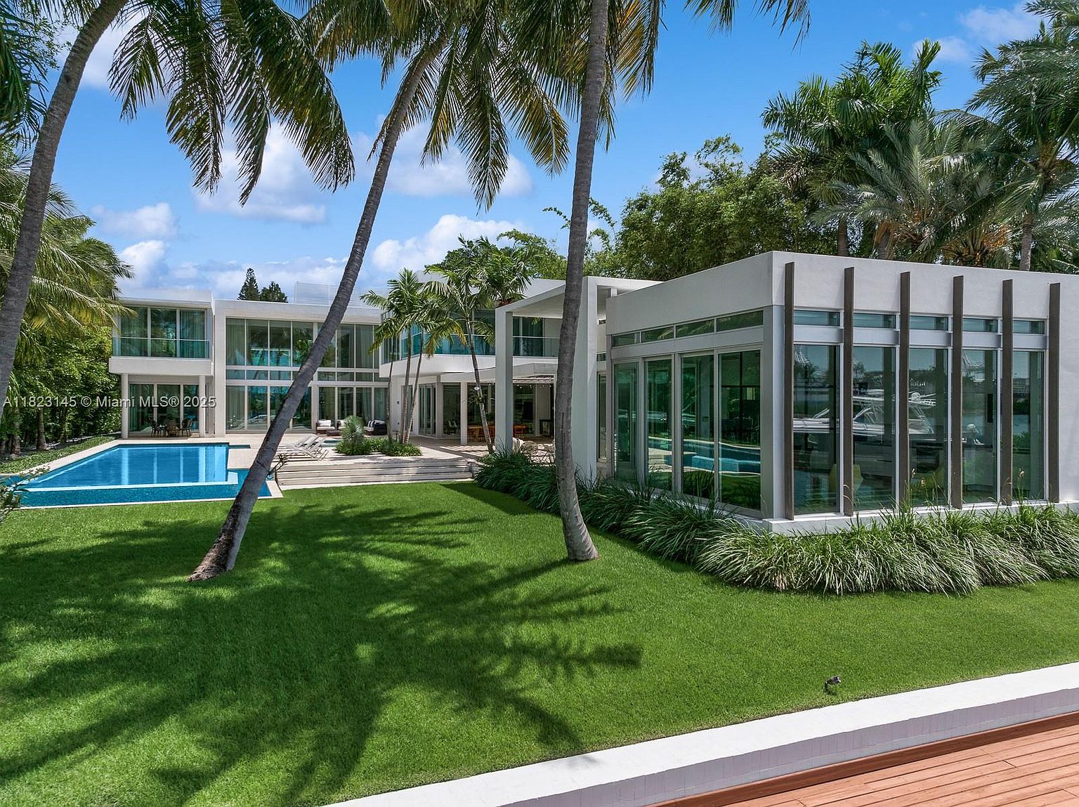 This modern luxury estate features a striking architectural design with extensive glass walls and clean, geometric lines. The property includes a pristine swimming pool set against a lush green lawn, framed by tall palm trees and mature landscaping. The perspective captures the seamless transition between the indoor living spaces and the expansive outdoor recreational area, emphasizing a sophisticated, resort-style atmosphere.
