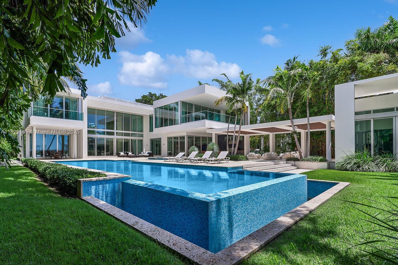 This modern luxury residence features a striking geometric design with extensive glass walls and a multi-level structure. A large, rectangular infinity-edge pool serves as the centerpiece of the backyard, complemented by a paved patio area with lounge chairs and a covered pergola. The lush green lawn and surrounding tropical landscaping create a serene, resort-like atmosphere.