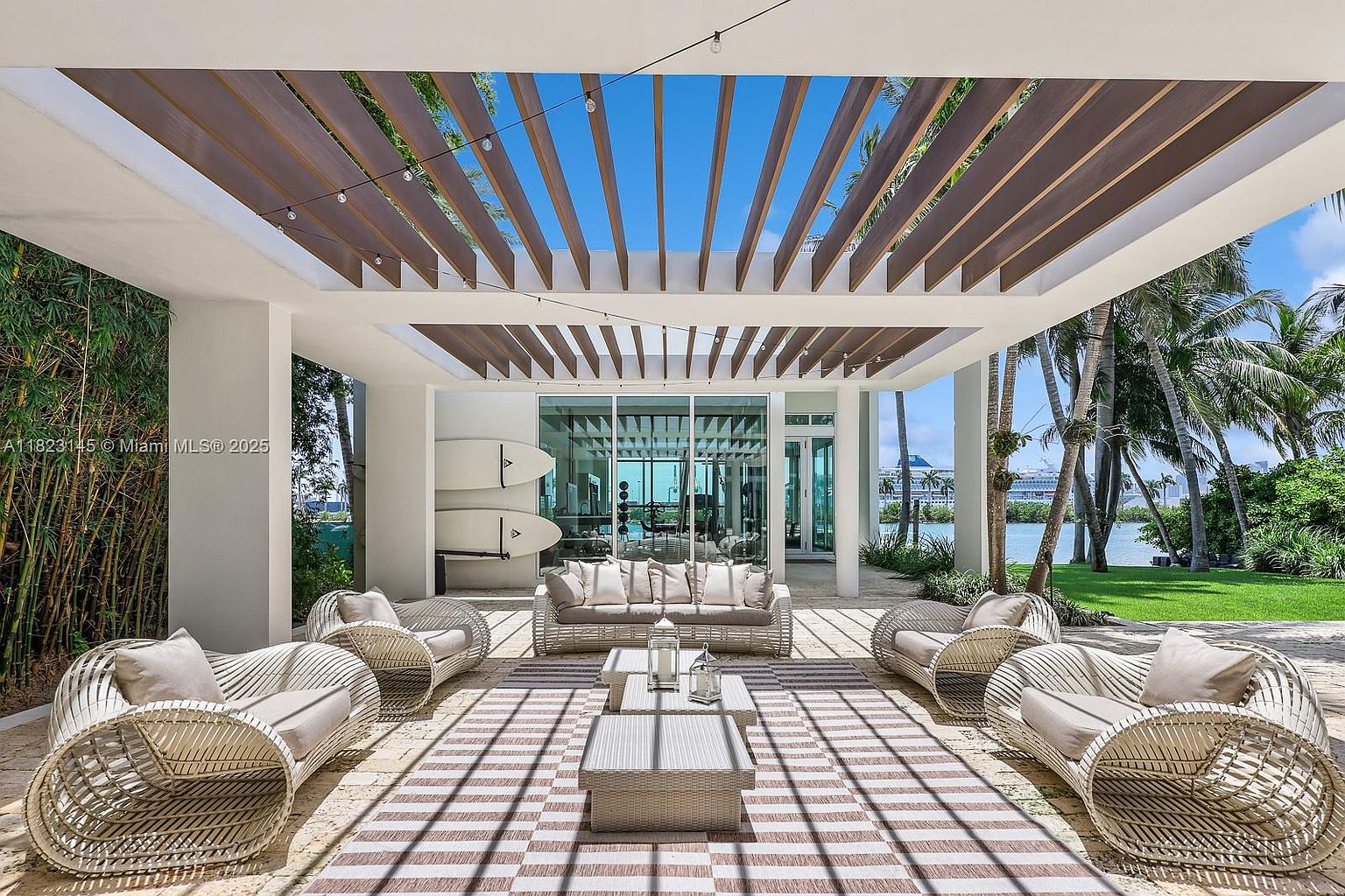 This expansive covered patio features a modern pergola roof structure that casts rhythmic shadows across the stone-paved floor. The space is furnished with light-toned, woven outdoor seating arranged around a central coffee table, creating a sophisticated lounge area. Beyond the seating, large glass doors provide a view into the home, while the open side reveals a lush green lawn, palm trees, and a waterfront vista.
