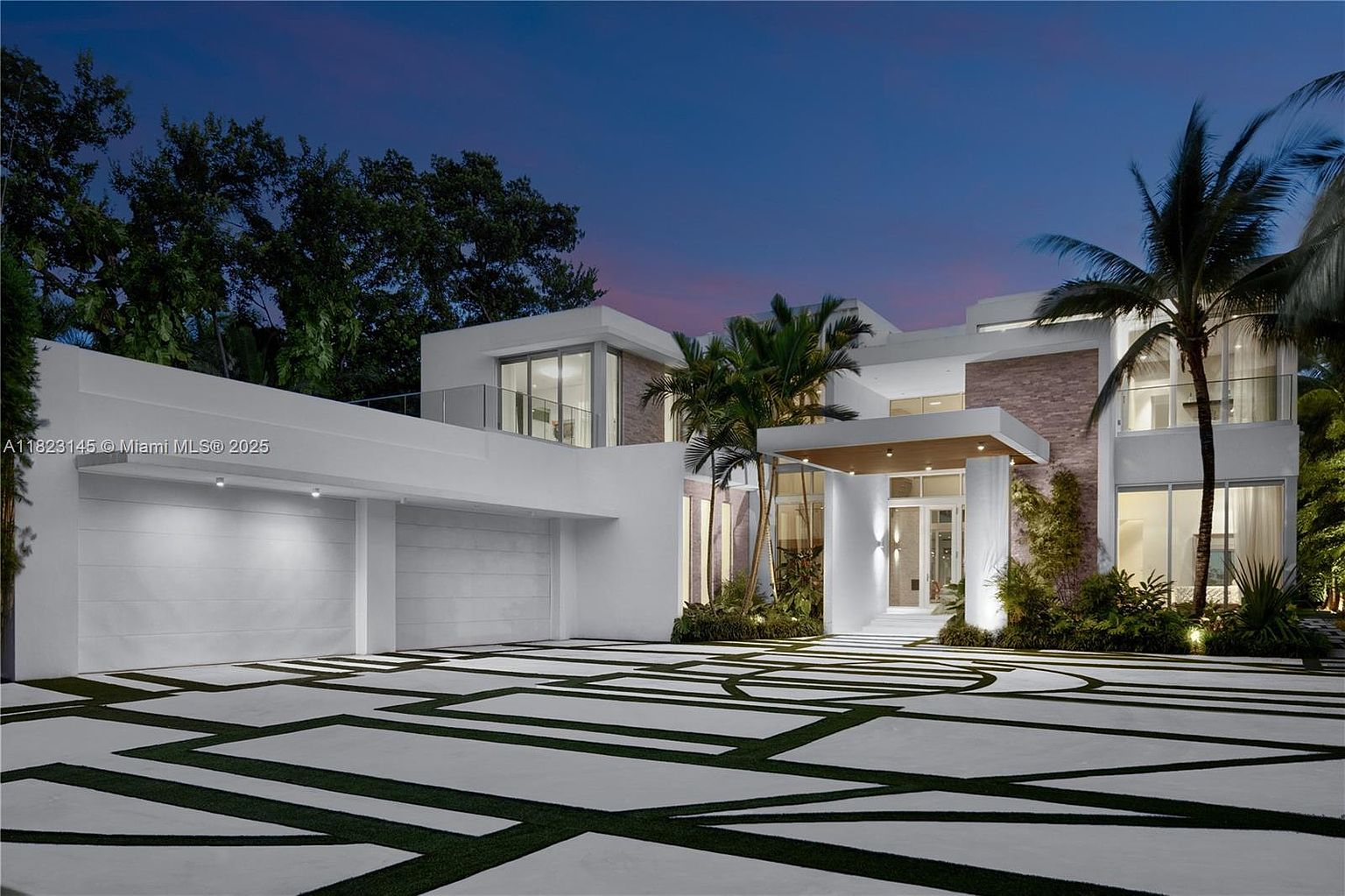 This striking modern residence features a clean, white geometric facade with expansive glass elements and a prominent covered entryway. The driveway is paved with a unique pattern of concrete slabs separated by dark, linear landscaping, leading to a multi-car garage on the left. Lush tropical palm trees and manicured greenery frame the structure, which is captured at dusk under a soft, twilight sky.