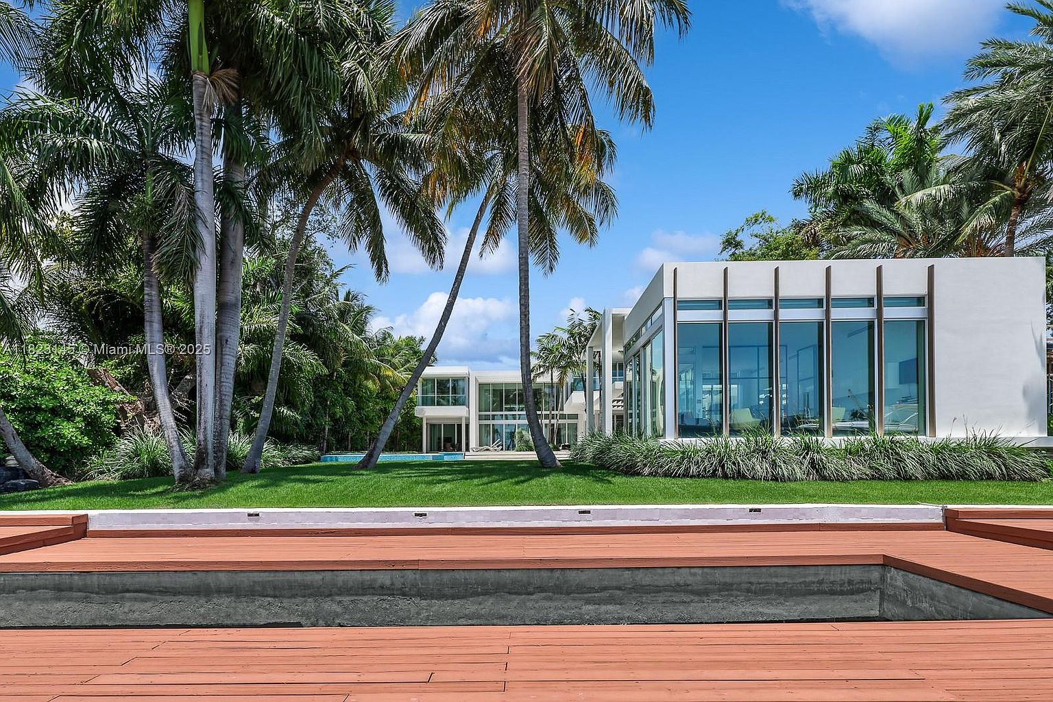 This rear view of a modern luxury estate showcases a sleek, white architectural design with extensive floor-to-ceiling glass walls that reflect the bright blue sky. A lush green lawn separates the residence from a foreground wooden deck, which features a recessed pool area. Tall palm trees frame the structure, creating a serene and tropical atmosphere typical of high-end coastal properties.