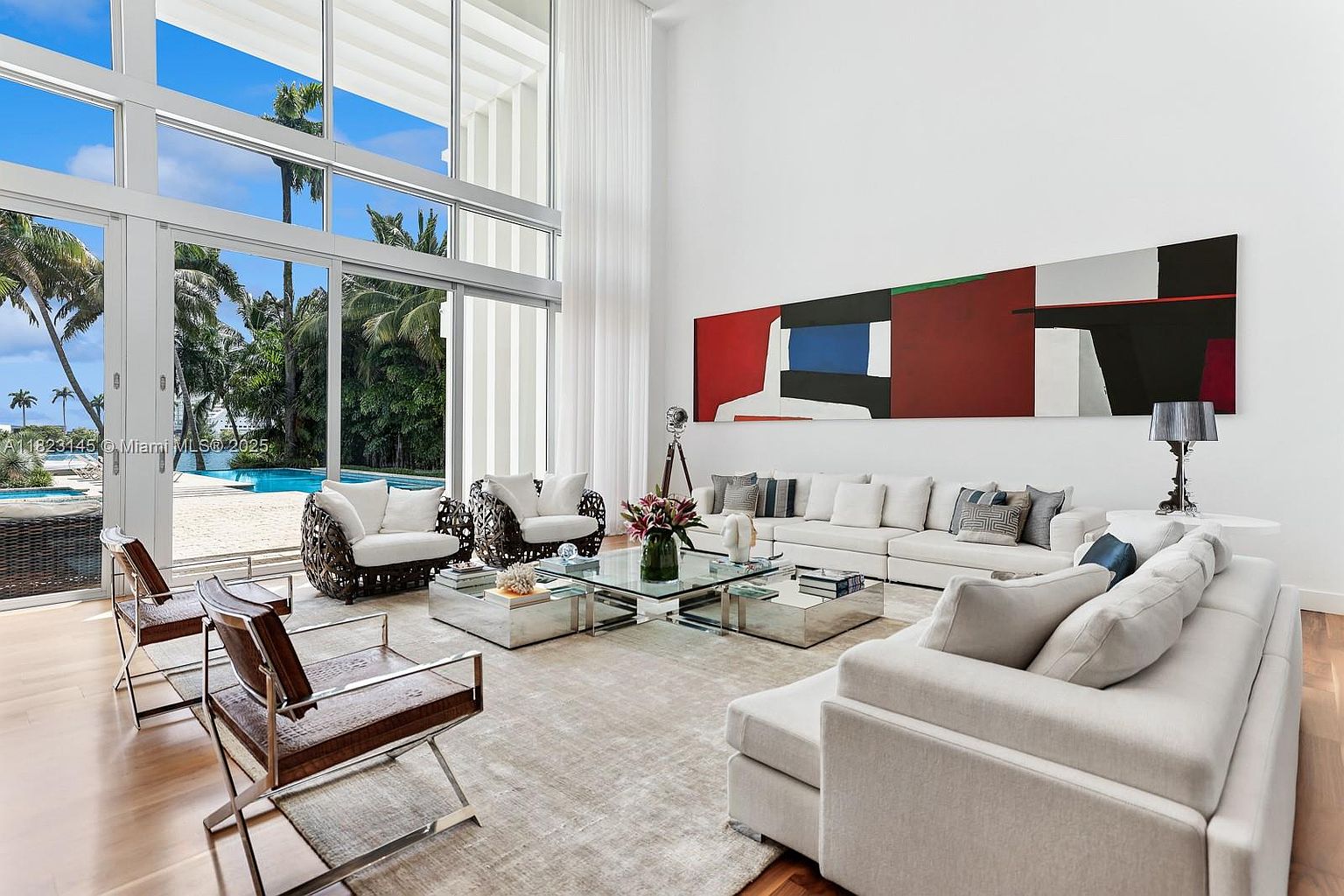 This expansive, double-height living room features a bright, airy aesthetic with large glass walls that provide a seamless view of the outdoor pool and lush tropical landscaping. The space is anchored by a large, neutral-toned sectional sofa and a central glass coffee table, complemented by modern accent chairs and a bold, abstract geometric painting on the wall. The room is bathed in natural light, creating a sophisticated and luxurious atmosphere perfect for entertaining.