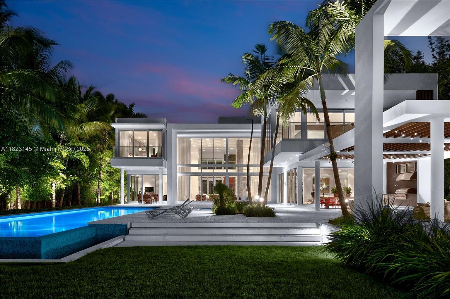 This stunning modern residence is captured at twilight, showcasing a dramatic rear facade with expansive glass walls that illuminate the interior. The property features a sleek, multi-level architectural design, a pristine swimming pool with integrated lighting, and a spacious stone patio perfect for outdoor entertaining. Lush tropical landscaping and tall palm trees frame the structure, creating a sophisticated and serene atmosphere.