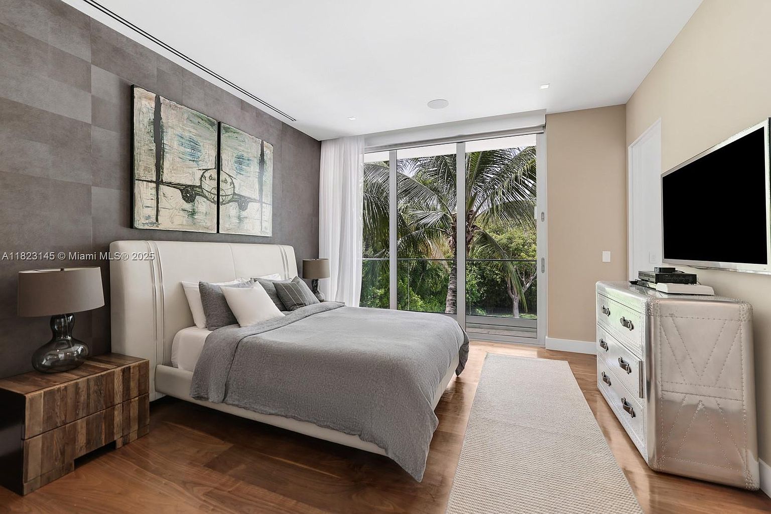 This primary bedroom features a contemporary aesthetic with a large, upholstered bed centered against a textured, dark-toned accent wall adorned with a diptych airplane-themed artwork. A metallic, industrial-style dresser sits to the right, while a warm-toned wood nightstand with a modern lamp anchors the left side. Large glass sliding doors provide a seamless transition to a balcony, offering a bright, airy view of lush tropical greenery outside.