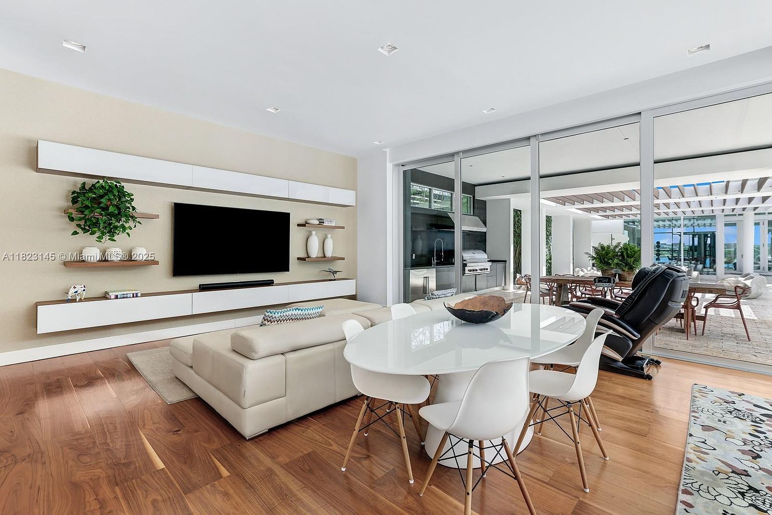 This open-concept living and dining area features warm wood flooring and a neutral color palette, creating a bright and inviting atmosphere. The living space is anchored by a wall-mounted television above a sleek, minimalist media console, while the adjacent dining area is furnished with a modern white oval table and matching chairs. Large glass sliding doors provide a seamless transition to an outdoor patio, enhancing the sense of space and natural light throughout the room.