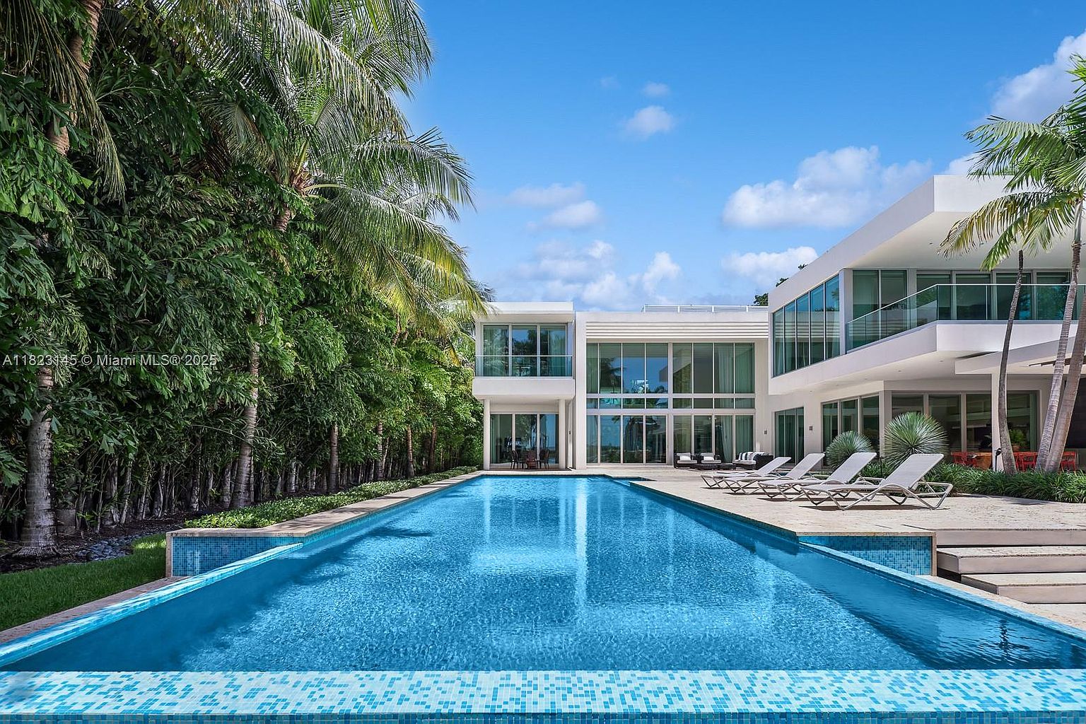 This modern luxury residence features a long, rectangular swimming pool as the centerpiece of the backyard, framed by a stone patio and a row of tall palm trees. The house showcases a contemporary architectural style with extensive glass walls and clean, white lines that reflect in the pool's surface. The perspective is a low-angle, eye-level shot looking directly toward the rear facade of the home, emphasizing the seamless transition between the indoor and outdoor living spaces.