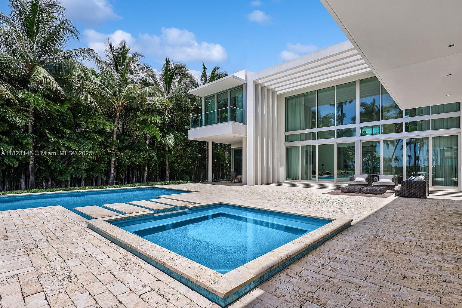 This modern luxury residence features a sprawling outdoor living area centered around a geometric swimming pool and integrated spa. The home showcases expansive glass walls that provide a seamless transition between the interior and the stone-paved patio, which is furnished with contemporary outdoor seating. Lush tropical landscaping and tall palm trees frame the property, creating a private and serene resort-like atmosphere.