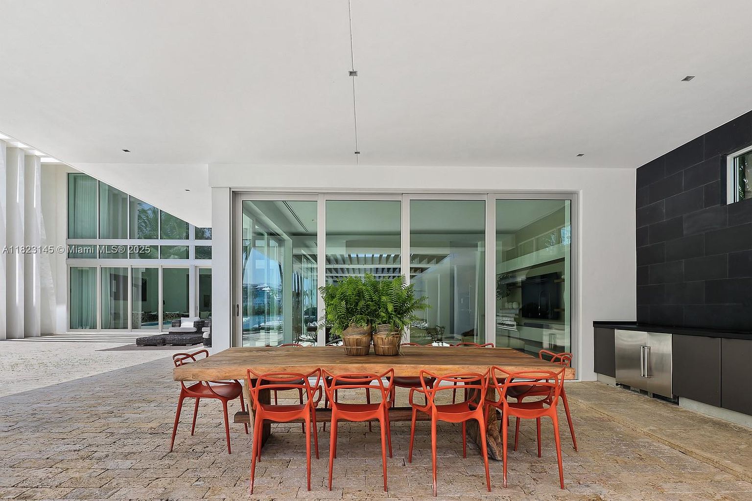 This expansive covered patio features a large, rustic wooden dining table surrounded by vibrant orange chairs, creating a striking contrast against the neutral stone flooring. The space is framed by floor-to-ceiling glass walls that provide a seamless transition to the interior living areas and a view of the outdoor pool beyond. A sleek, dark-toned outdoor kitchen unit with stainless steel appliances is positioned to the side, enhancing the functionality of this sophisticated al fresco dining area.