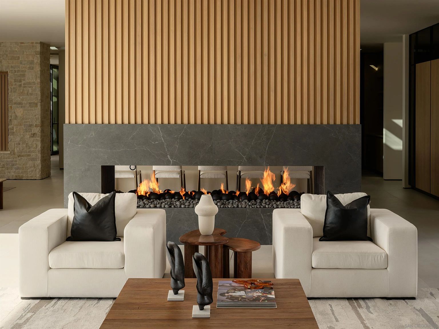 This sophisticated living space centers on a double-sided fireplace featuring a dark stone surround and a dramatic vertical wood-slat wall above. Two plush, cream-colored armchairs with black leather accent pillows flank a small wooden side table, while a larger wooden coffee table sits in the foreground. The room exudes a modern, minimalist aesthetic with a neutral color palette and clean, symmetrical lines.