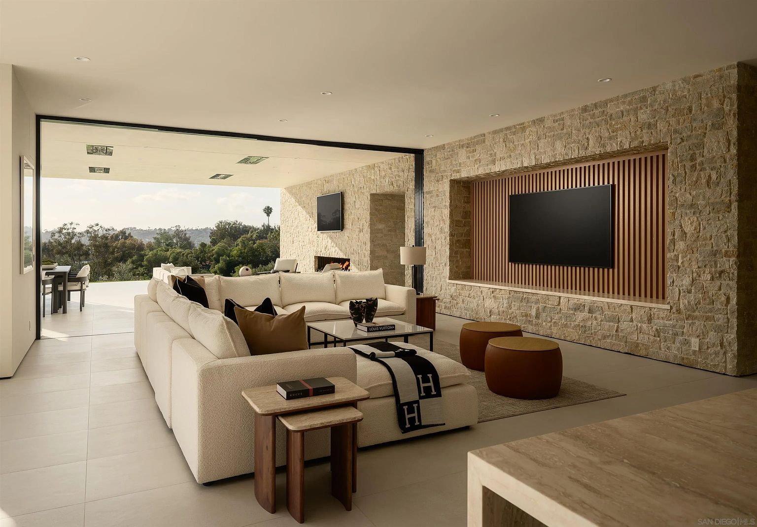 This spacious, modern living room features a large, light-colored sectional sofa and a central coffee table, creating an inviting seating area. A prominent stone-clad wall serves as a focal point, housing a large television set against a backdrop of vertical wood slats. The room seamlessly transitions to an expansive outdoor patio through wide, open glass panels, offering a panoramic view of the surrounding landscape.