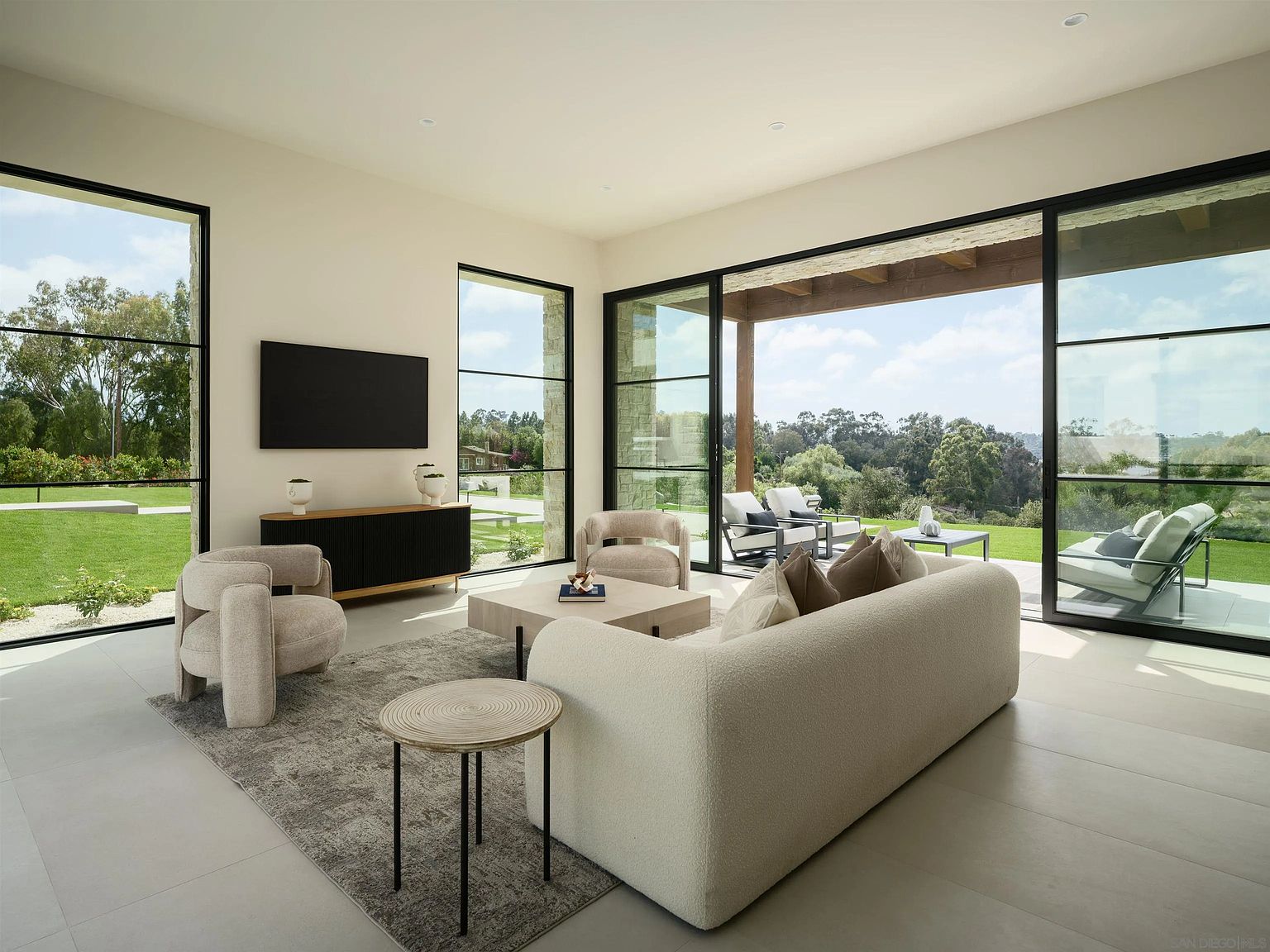 This bright and airy living room features a contemporary design with a neutral color palette and plush, curved furniture. Large, floor-to-ceiling glass sliding doors open to a covered patio, seamlessly blending the indoor space with the lush, green landscape outside. The room is anchored by a textured area rug and a minimalist media console, creating a sophisticated and inviting atmosphere.