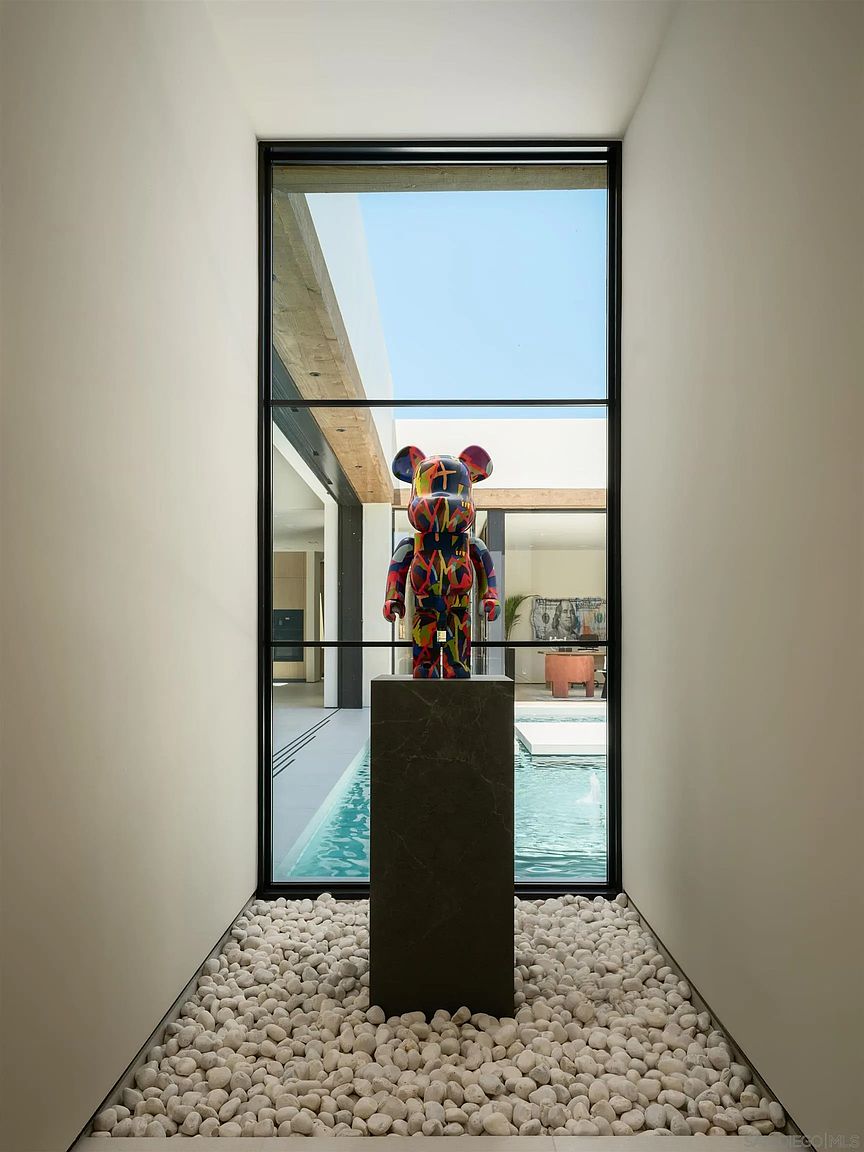 This striking interior hallway features a minimalist design centered around a large, floor-to-ceiling window that frames a view of a courtyard pool. A colorful, modern art sculpture stands prominently on a dark stone pedestal, set against a bed of white decorative river stones. The perspective is symmetrical and cinematic, drawing the eye through the glass to the outdoor water feature and the bright, airy architecture beyond.