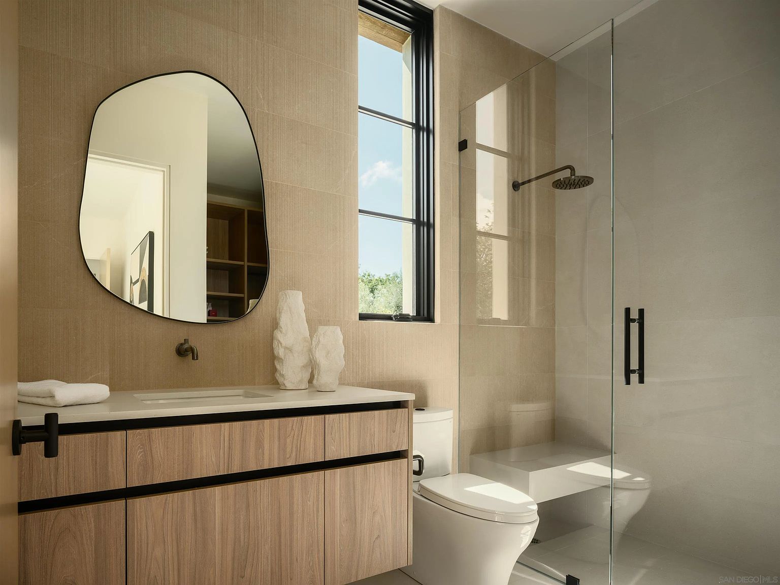 This modern bathroom features a light wood vanity with a clean-lined countertop and a unique, organic-shaped mirror. A glass-enclosed shower with a built-in bench sits adjacent to a sleek toilet, all set against textured, neutral-toned wall tiles. The space is illuminated by natural light from a tall, narrow window, creating a bright and contemporary atmosphere.