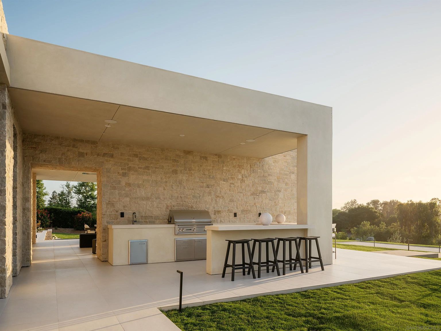 This sophisticated outdoor living space features a covered patio with a built-in kitchen, complete with a stainless steel grill and a stone-clad wall. A sleek bar counter with four stools provides an ideal spot for entertaining, while the expansive, light-toned tiled flooring extends out toward a manicured lawn. The architectural design emphasizes clean lines and a seamless transition between the indoor-outdoor living areas, set against a serene, sun-drenched landscape.