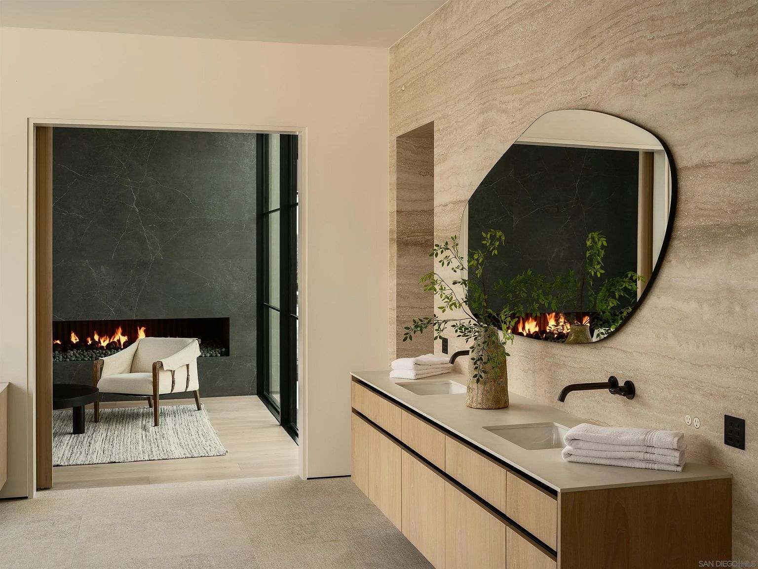 This sophisticated primary bathroom features a dual-vanity with light wood cabinetry and a smooth, neutral-toned countertop. An organic-shaped mirror reflects a view into an adjacent lounge area, which is anchored by a dark stone fireplace and a comfortable armchair. The space is defined by its clean lines, high-end finishes, and a seamless transition between the functional bathroom area and the relaxing fireplace lounge.