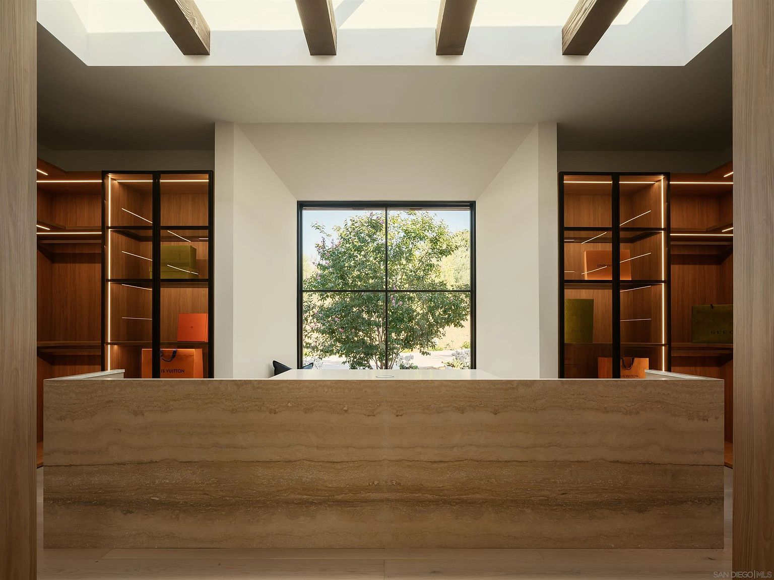 This sophisticated walk-in closet features a central island with a prominent stone countertop and a warm, layered texture. Flanking the island are symmetrical, illuminated shelving units with glass doors, showcasing designer shopping bags and providing a boutique-like display. The space is bathed in natural light from a large, grid-paned window that frames a view of the outdoors, while exposed wooden ceiling beams add architectural character to the room.