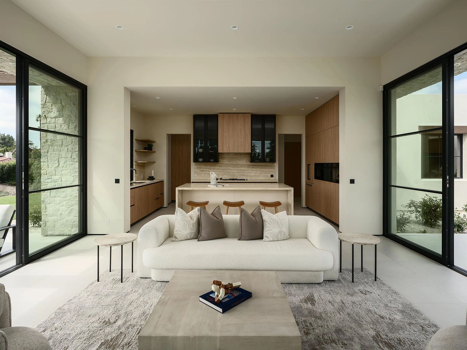 This open-concept living space features a contemporary white sofa centered on a textured area rug, facing a minimalist light wood coffee table. The room is framed by expansive floor-to-ceiling glass walls on both sides, providing a seamless transition to the outdoor environment. Beyond the living area, a sleek kitchen with light wood cabinetry, a stone-topped island, and dark accent cabinets creates a sophisticated, modern aesthetic.