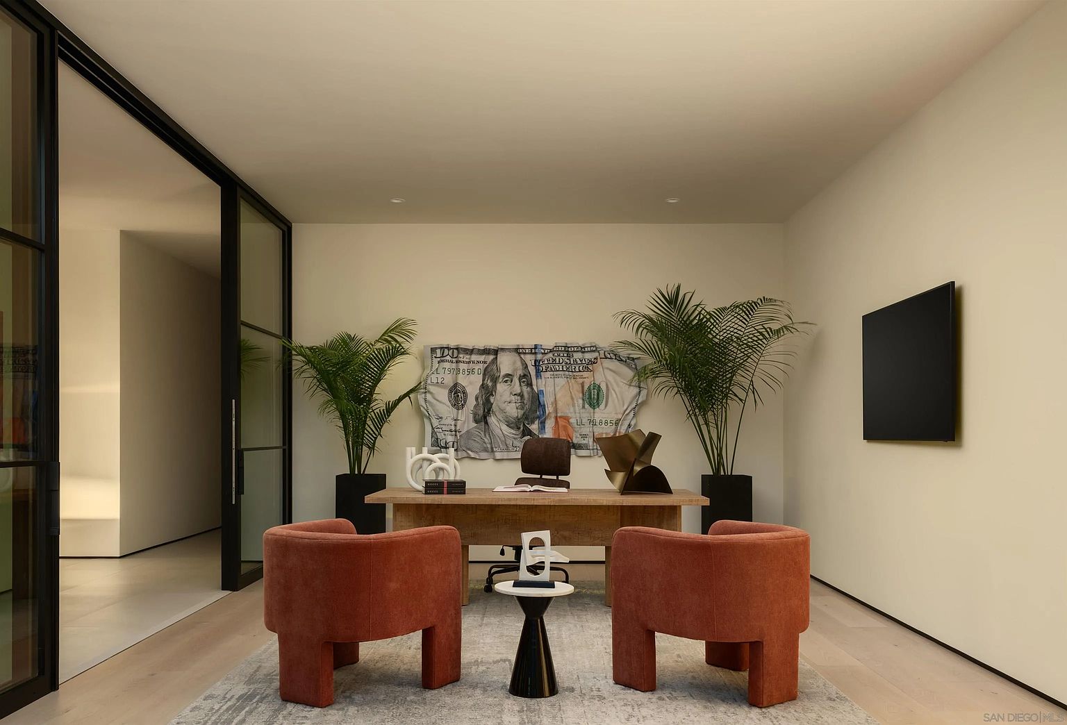 This modern home office features a minimalist aesthetic with a light wood desk positioned centrally, flanked by two vibrant orange armchairs. A large, artistic print of a hundred-dollar bill serves as a bold focal point on the back wall, complemented by two potted palm plants. The room is framed by sleek black-trimmed glass doors and light-toned flooring, creating a sophisticated and professional workspace.