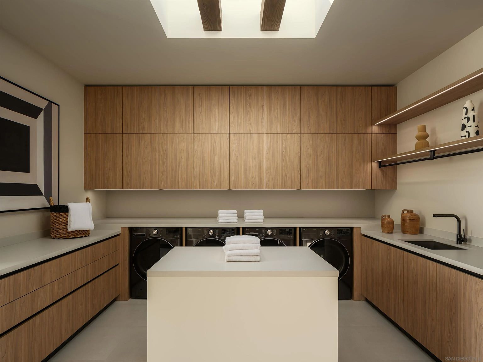This modern, symmetrical laundry room features sleek light wood cabinetry and a central island with a light-colored countertop. The space is equipped with four side-by-side black appliances, a built-in sink, and open shelving, all illuminated by a skylight above. The clean lines and neutral color palette create a highly organized and functional atmosphere.
