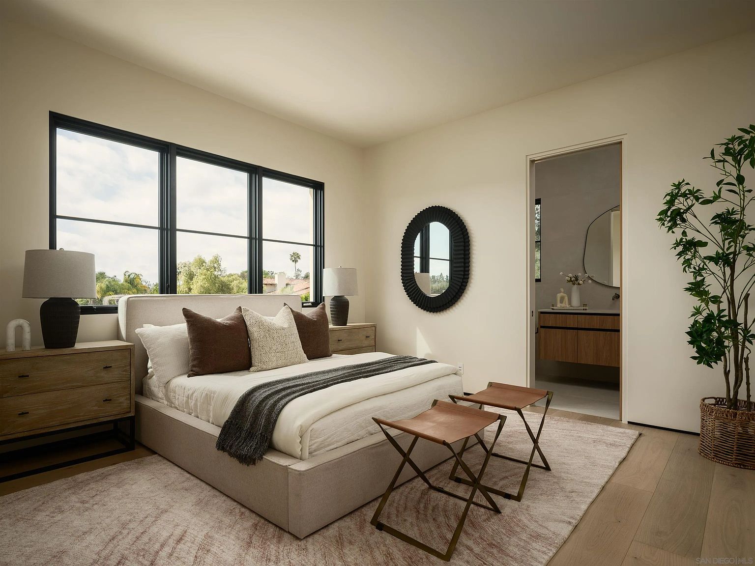 This serene primary bedroom features a neutral color palette with a plush upholstered bed frame and layered bedding in shades of cream and brown. Large windows with black frames allow for ample natural light, while a decorative oval mirror and two leather-topped stools add a touch of modern sophistication. The room flows seamlessly into an adjacent bathroom, creating an open and airy atmosphere.