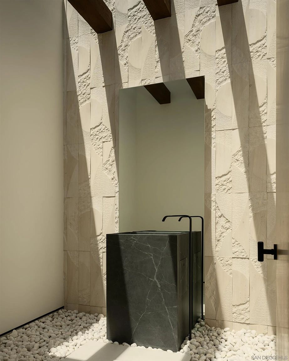 This minimalist powder room features a striking dark stone pedestal sink paired with a sleek, black wall-mounted faucet. The space is defined by a textured, light-colored stone wall that creates a dramatic interplay of light and shadow, complemented by a bed of white river stones on the floor. A tall, frameless mirror is integrated into the stone wall, enhancing the clean, contemporary aesthetic of this unique architectural space.