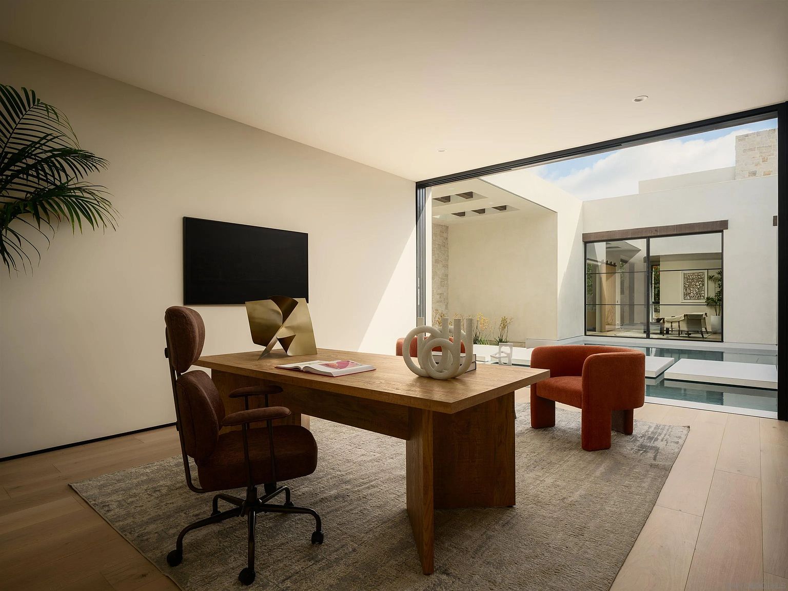 This sophisticated home office features a minimalist design with a large wooden desk as the central focal point, complemented by a brown ergonomic chair and a burnt-orange accent chair. The room is bathed in natural light from expansive floor-to-ceiling glass walls that provide a seamless transition to an outdoor courtyard with a swimming pool. The neutral color palette and clean lines create a serene, professional atmosphere ideal for focused work.