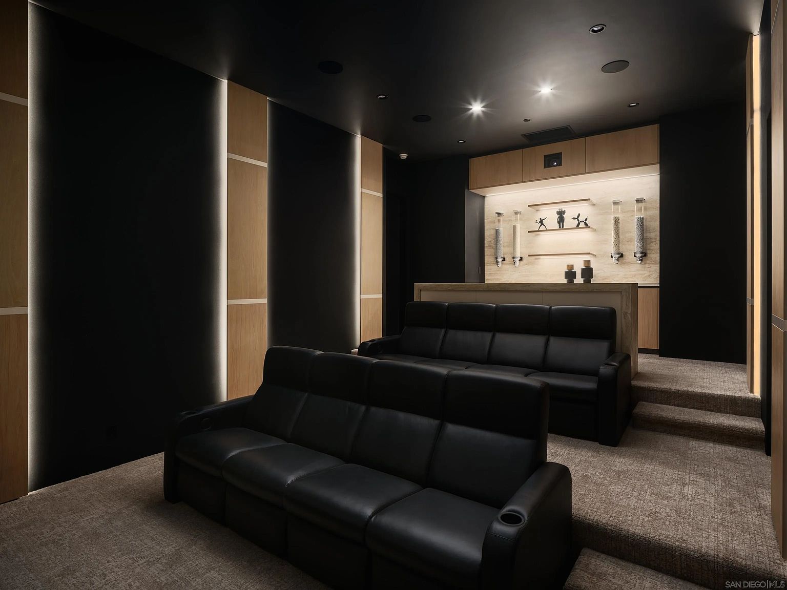 This sophisticated home theater features two rows of tiered black leather reclining seating set against a dark, moody color palette. The room is accented with vertical light wood panels illuminated by integrated LED strips, creating a modern and cinematic atmosphere. At the rear, a built-in refreshment bar includes shelving with decorative figurines and wall-mounted dispensers, providing a functional and stylish entertainment space.