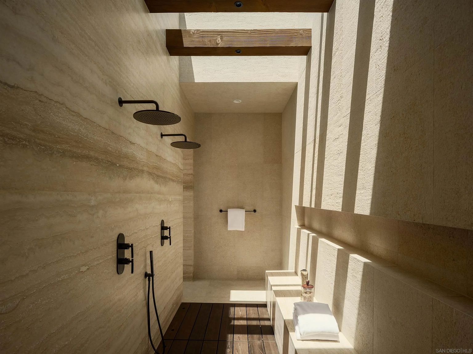 This luxurious walk-in shower features a striking combination of textured stone walls and warm wood-slat flooring. The space is illuminated by natural light streaming through overhead wooden beams, creating dramatic linear shadows across the stone surfaces. It includes dual shower heads, matte black fixtures, a built-in stone bench, and a minimalist towel rack, offering a spa-like atmosphere.