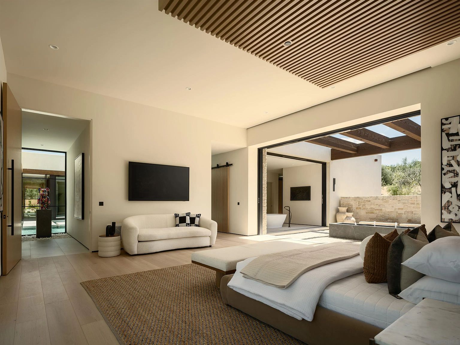 This expansive primary bedroom features a seamless transition to an outdoor patio through large sliding glass doors, creating an indoor-outdoor living experience. The room is anchored by a neutral-toned bed and a textured area rug, complemented by a minimalist sofa and a wall-mounted television. A slatted wood ceiling detail adds architectural interest, while the open-concept design allows for a view into an adjacent bathroom area with a soaking tub.