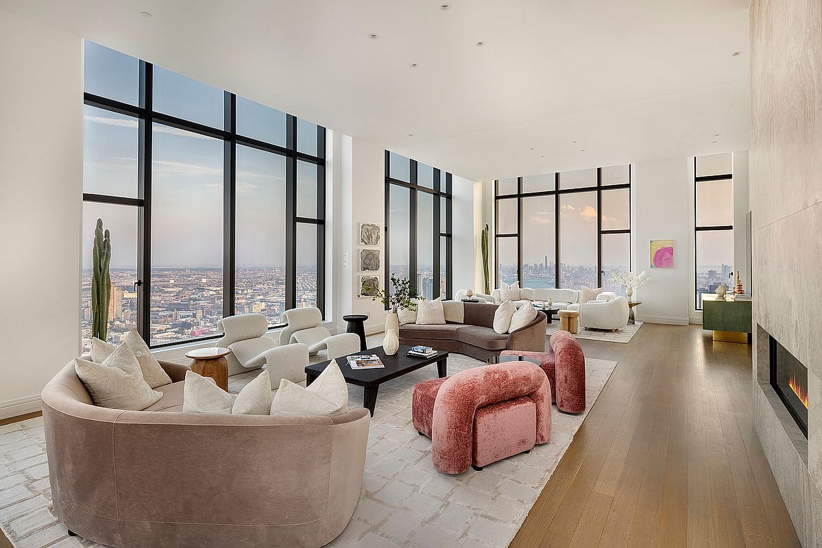 This expansive, high-end living room features floor-to-ceiling windows that offer a breathtaking panoramic view of the city skyline. The space is elegantly furnished with a mix of plush, curved sofas and modern armchairs in soft, neutral and dusty rose tones, arranged on a large, textured area rug. A sleek fireplace integrated into a stone wall adds a sophisticated focal point, creating an atmosphere of refined luxury and comfort.