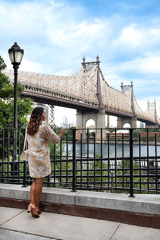 This image captures a scenic waterfront promenade featuring a woman standing by a black metal railing, looking out toward the iconic Queensboro Bridge. The setting highlights a desirable urban lifestyle with easy access to landmark views and public outdoor spaces. The composition emphasizes the bridge's intricate steel architecture against a bright, cloudy sky, creating an inviting and picturesque atmosphere.