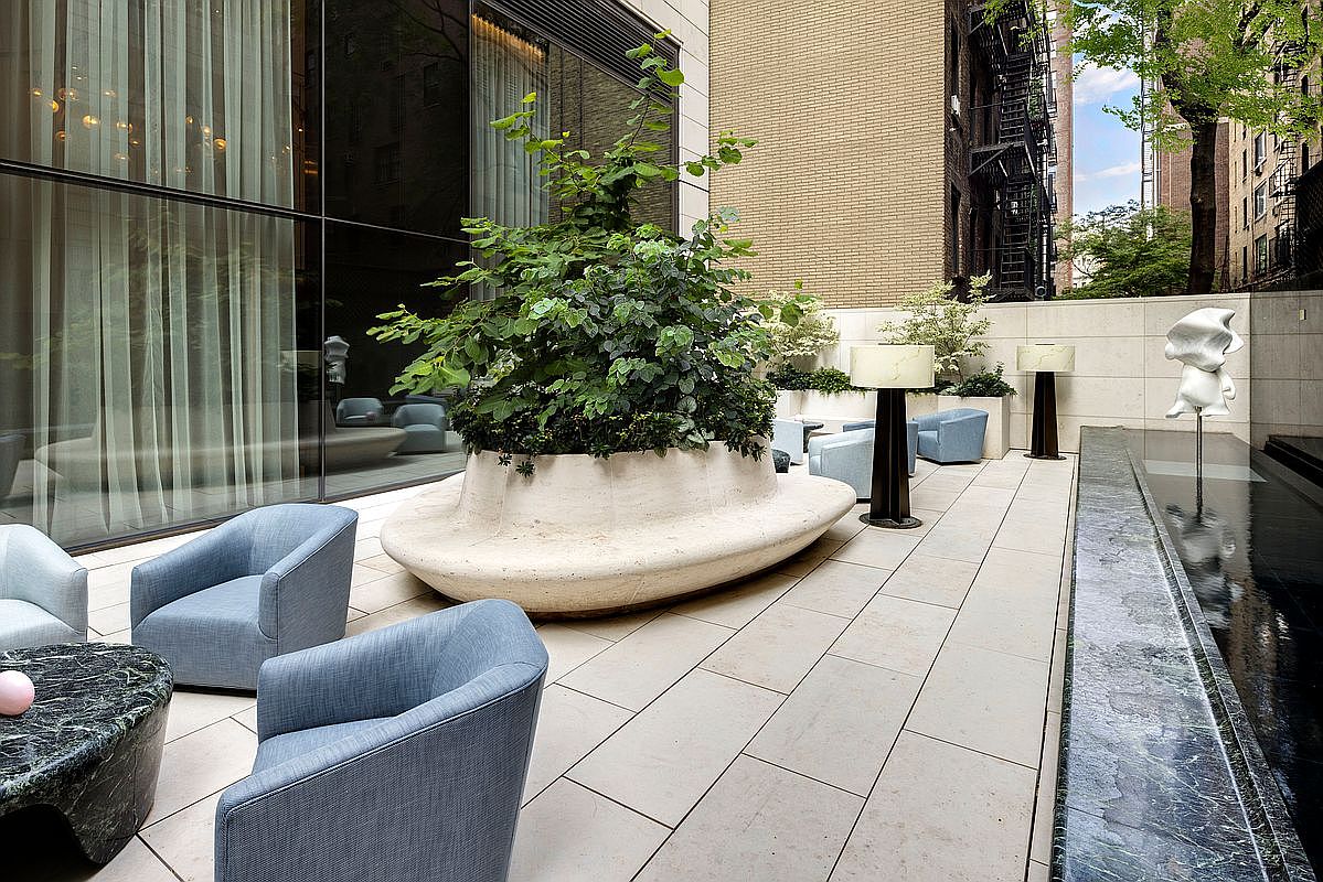 This sophisticated urban terrace features a sleek, modern design with large stone pavers and a central sculptural planter housing a lush green tree. The space is furnished with contemporary blue armchairs and a marble-topped coffee table, complemented by a serene water feature and an abstract white sculpture. The cinematic perspective captures a tranquil, high-end outdoor lounge area that seamlessly blends architectural elegance with natural elements.