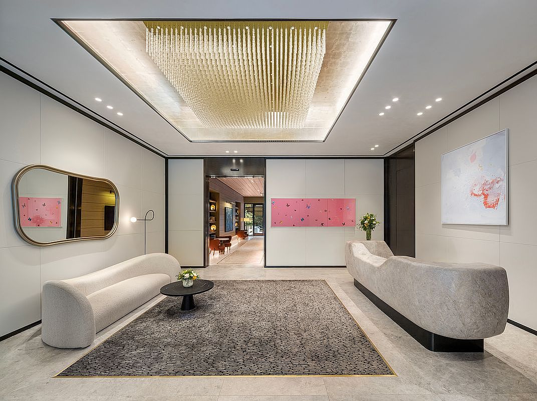 This sophisticated lobby or entryway features a grand, tiered crystal chandelier suspended from a recessed ceiling, casting a warm glow over the space. The room is furnished with a curved, cream-colored sofa, a minimalist black coffee table, and a substantial stone-textured reception desk, all set against a backdrop of neutral walls adorned with contemporary art. The perspective is a wide, symmetrical shot that emphasizes the clean lines, luxurious materials, and elegant, modern aesthetic of the building's entrance.