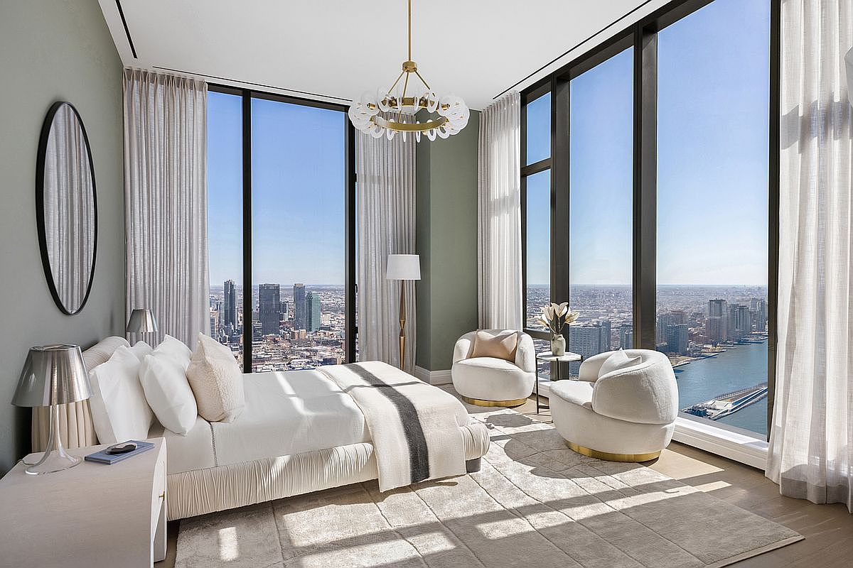 This luxurious primary bedroom features floor-to-ceiling windows that offer a breathtaking, panoramic view of the city skyline and river. The room is elegantly furnished with a plush bed, two curved cream-colored armchairs, and a sophisticated chandelier, all set against a soft, neutral color palette. The high-end design creates an airy, serene atmosphere that emphasizes the spectacular urban vista.