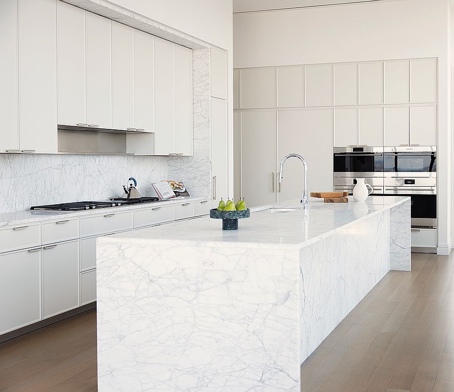 This sleek, high-end kitchen features a massive waterfall-edge marble island as its centerpiece, complemented by clean white cabinetry and integrated stainless steel appliances. The minimalist design is enhanced by a matching marble backsplash and light hardwood flooring, creating a bright and sophisticated atmosphere. The perspective is a wide-angle shot that emphasizes the expansive, open-concept layout and the luxurious materials used throughout the space.