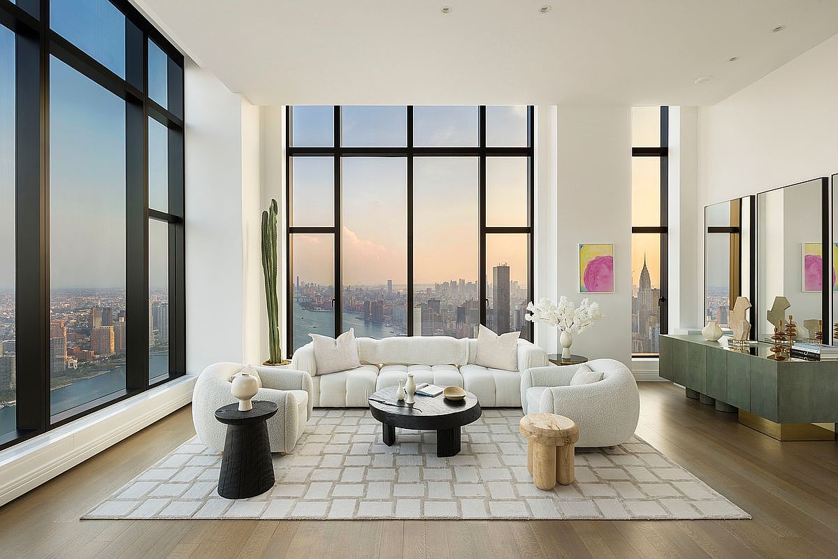 This sophisticated, high-rise living room features floor-to-ceiling windows that offer a breathtaking, panoramic view of the New York City skyline and river. The space is furnished with a plush, cream-colored curved sofa and matching armchairs arranged on a textured area rug, centered around a dark, minimalist coffee table. A sleek, olive-green credenza sits against the wall, complemented by modern art and sculptural decor, creating an atmosphere of refined, contemporary luxury.