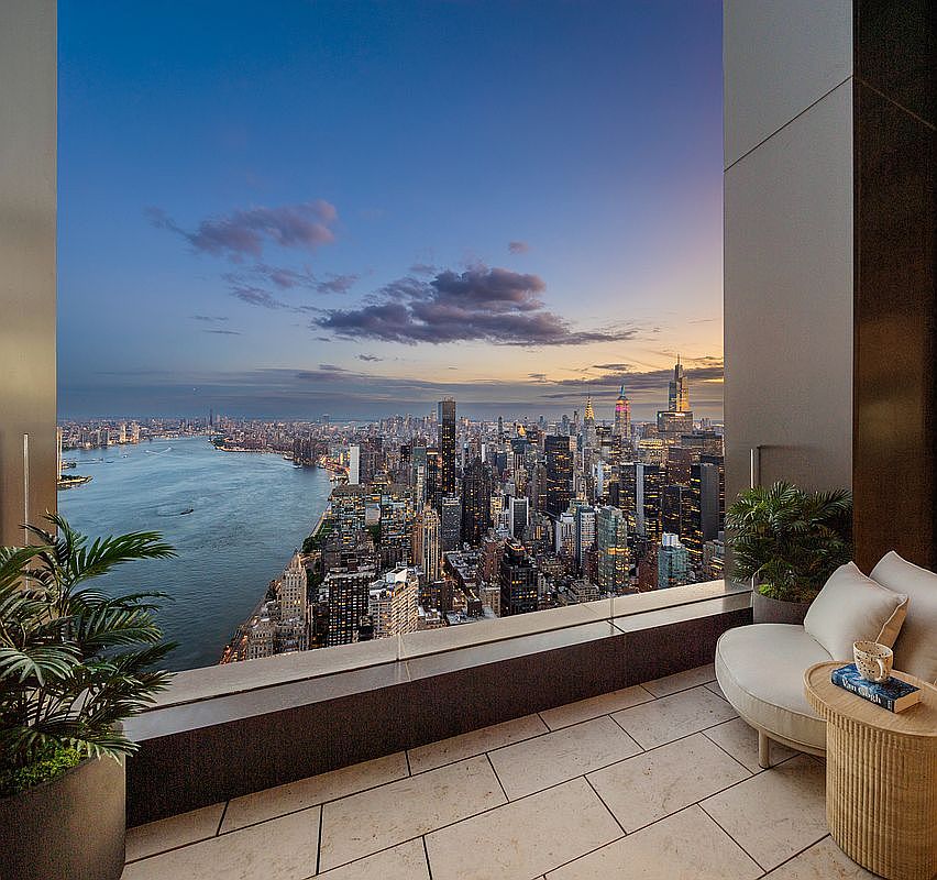 This luxurious high-rise balcony offers a breathtaking, panoramic view of the New York City skyline and the Hudson River at dusk. The space is elegantly furnished with a comfortable, curved lounge chair, a textured side table, and lush potted plants, creating a serene and sophisticated outdoor retreat. The perspective is cinematic, framing the vast urban landscape through the clean lines of the modern architectural structure.
