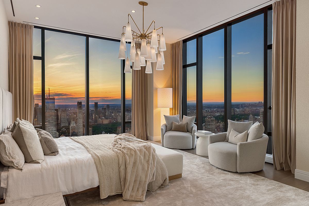 This luxurious primary bedroom features floor-to-ceiling windows that offer a breathtaking, panoramic sunset view of the city skyline and Central Park. The room is elegantly furnished with a plush, neutral-toned bed, a cozy seating area with two swivel chairs, and a sophisticated modern chandelier. The overall aesthetic is one of high-end, serene urban living, emphasizing comfort and spectacular natural light.