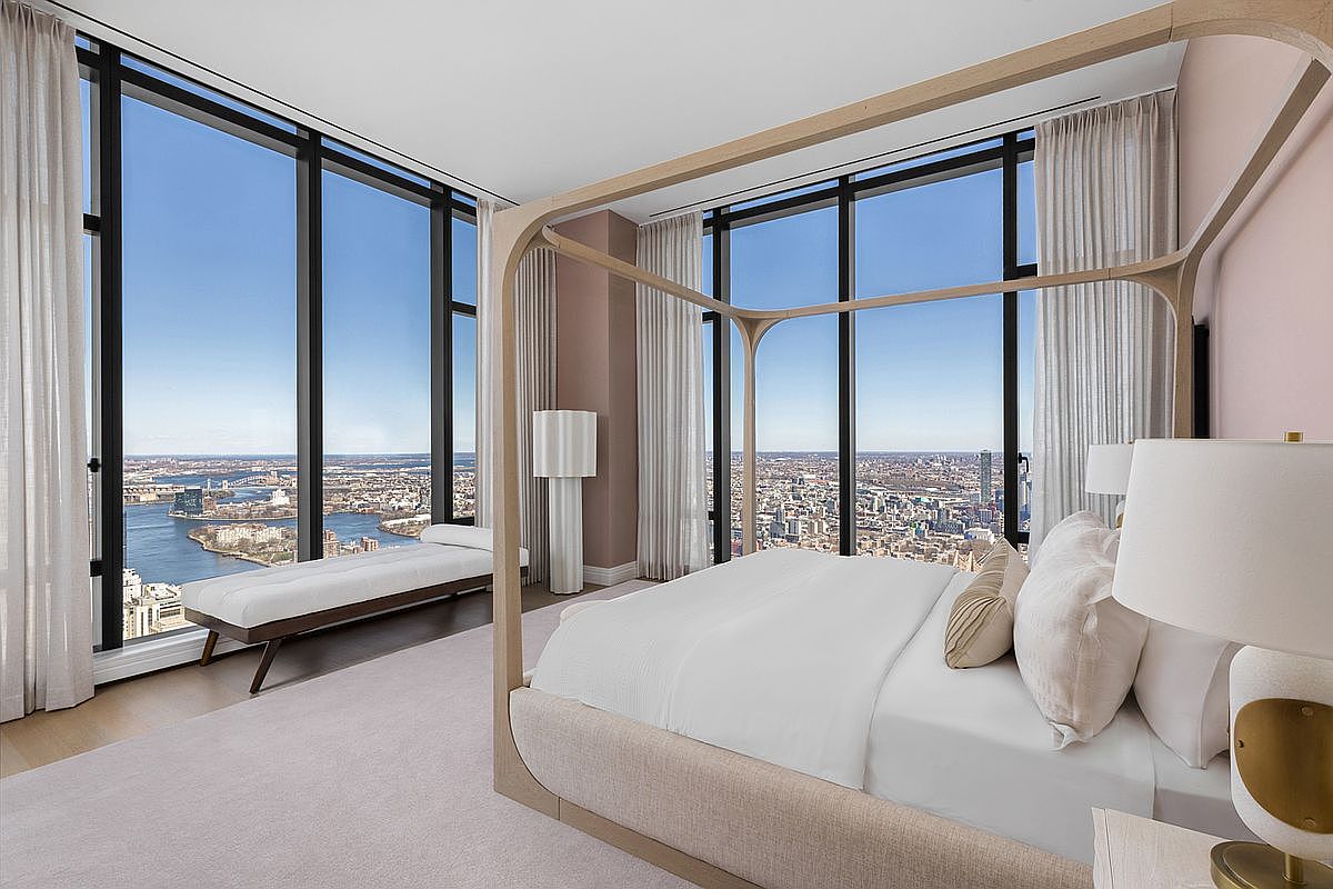 This luxurious primary bedroom features a modern four-poster wooden bed frame as the centerpiece, set against floor-to-ceiling windows that offer a breathtaking panoramic view of the city skyline and river. The room is decorated in a soft, neutral palette with plush carpeting and elegant sheer curtains, creating a serene and sophisticated atmosphere. A sleek bench sits by the window, providing a perfect vantage point to enjoy the expansive urban vista.