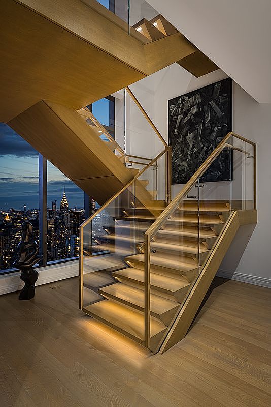 This striking interior shot features a modern, sculptural wooden staircase with integrated LED lighting and glass railings, serving as a dramatic focal point. The perspective captures the staircase from a low angle, emphasizing its architectural elegance against a backdrop of floor-to-ceiling windows that reveal a breathtaking nighttime view of the New York City skyline, including the Chrysler Building. A contemporary abstract painting and a minimalist sculpture complete the sophisticated, high-end aesthetic of this luxury residence.