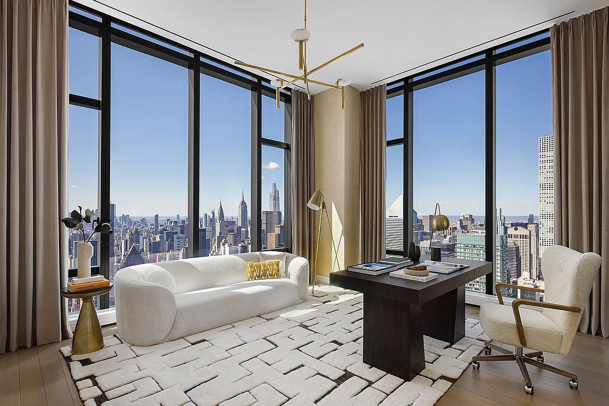 This sophisticated home office features floor-to-ceiling windows that offer a breathtaking, panoramic view of the New York City skyline. The room is furnished with a modern, curved white sofa, a sleek dark wood desk, and a textured area rug, creating a luxurious and professional atmosphere. The combination of high-end contemporary furniture and the dramatic urban backdrop makes this an ideal space for focused work or high-level meetings.