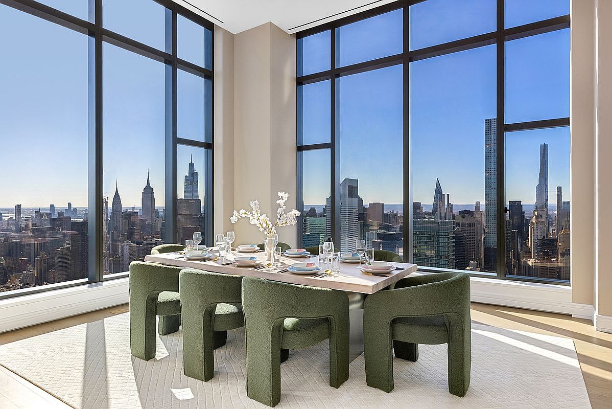 This sophisticated dining room features a modern, light-wood table surrounded by six plush, olive-green upholstered chairs, set against a backdrop of floor-to-ceiling windows. The expansive glass walls offer a breathtaking, panoramic view of the New York City skyline, creating a dramatic and luxurious atmosphere. The minimalist decor and neutral area rug emphasize the high-end, urban aesthetic of this high-rise residence.