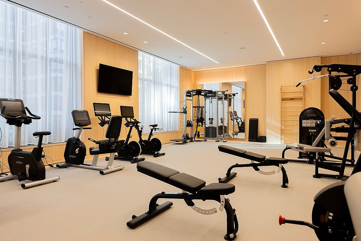 This modern, well-lit home gym features a variety of professional-grade exercise equipment, including stationary bikes, a multi-station cable machine, and adjustable weight benches. The space is characterized by light wood-paneled walls, neutral flooring, and floor-to-ceiling windows draped with sheer curtains that allow for ample natural light. The clean, minimalist aesthetic creates an inviting and functional environment for fitness enthusiasts.