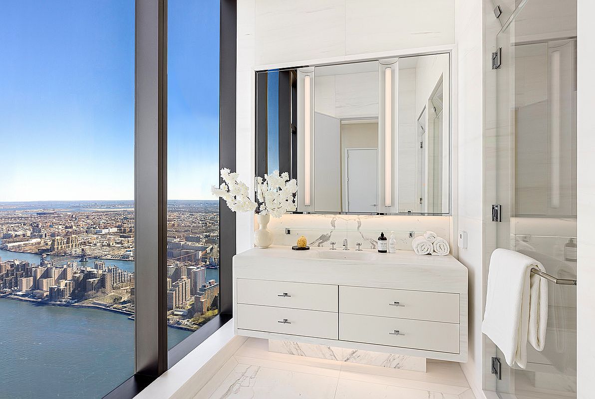 This luxurious primary bathroom features a sleek, floating white vanity with marble countertops and integrated lighting, set against a backdrop of floor-to-ceiling windows offering a breathtaking, high-altitude panoramic view of the city and river. The space is finished with elegant marble flooring and a glass-enclosed shower, creating a bright, sophisticated, and airy atmosphere. The combination of modern design and the dramatic urban vista provides a high-end, serene experience.