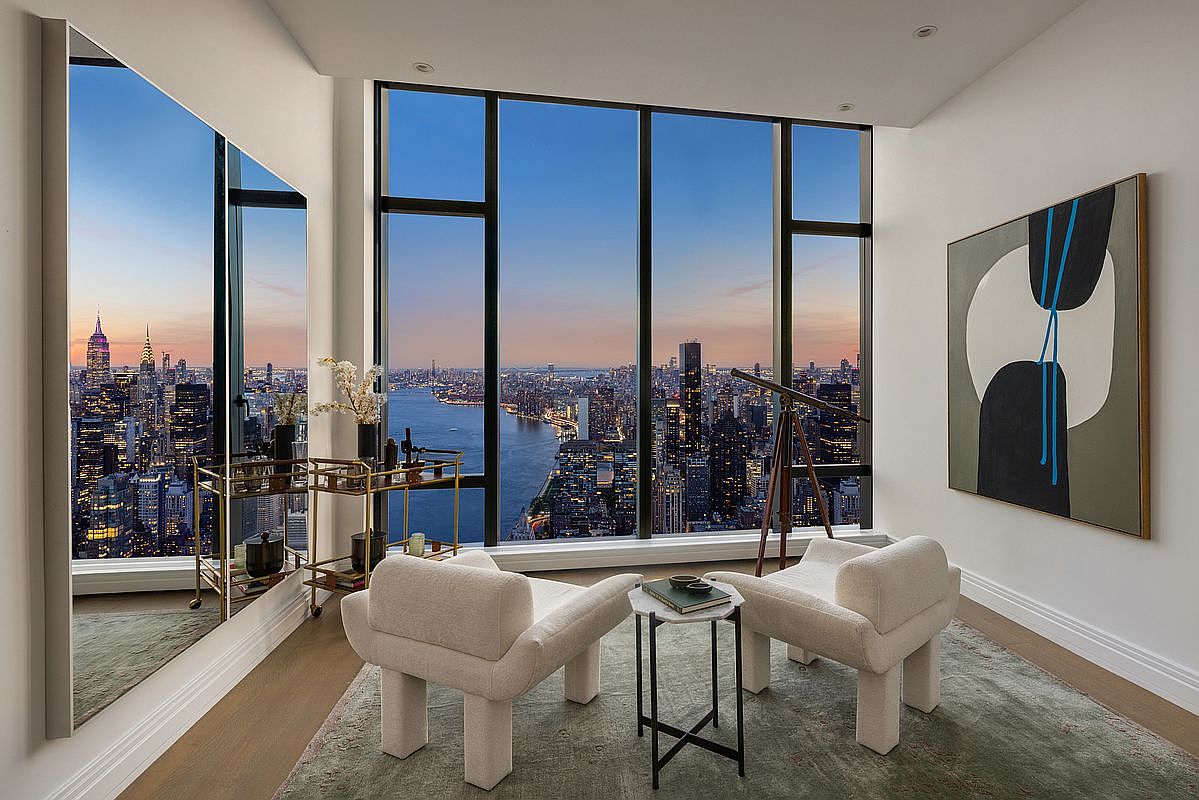 This sophisticated, high-rise lounge area features floor-to-ceiling windows that offer a breathtaking, panoramic view of the New York City skyline at dusk. The space is furnished with two plush, cream-colored armchairs, a sleek marble-topped side table, and a gold-accented bar cart, all set against a large, abstract piece of wall art. The combination of modern luxury furniture and the dramatic urban backdrop creates an atmosphere of refined elegance and exclusivity.