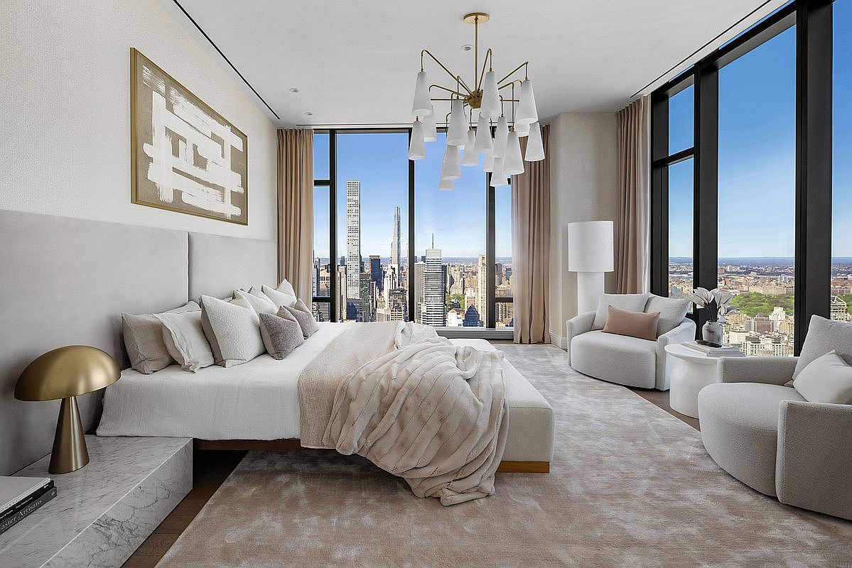This luxurious primary bedroom features floor-to-ceiling windows that offer a breathtaking, panoramic view of the New York City skyline. The room is elegantly appointed with a plush, neutral-toned color palette, a modern chandelier, and high-end marble accents, creating a sophisticated and serene atmosphere. The perspective is wide and inviting, showcasing the seamless blend of contemporary interior design and iconic urban scenery.