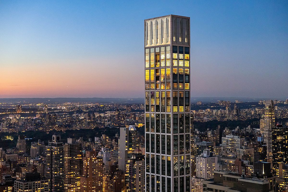 This striking aerial view captures a slender, ultra-luxury skyscraper rising prominently above the dense, illuminated cityscape of New York City at twilight. The building features a distinctive grid-like facade with warm, glowing interior lights that contrast beautifully against the deepening blue sky. The perspective emphasizes the tower's immense height and architectural elegance, showcasing its dominance over the surrounding urban skyline.