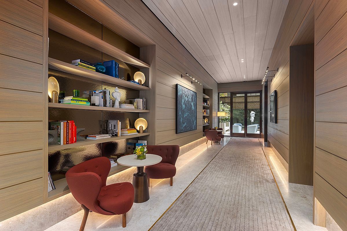 This sophisticated hallway features warm wood-paneled walls, recessed floor lighting, and a long, textured runner rug that leads toward a glass-paned entryway. A cozy seating area with two red armchairs and a small round table is nestled against a built-in bookshelf, creating an inviting and luxurious atmosphere. The perspective is a wide-angle shot looking down the corridor, emphasizing the clean lines, modern art, and elegant architectural details.